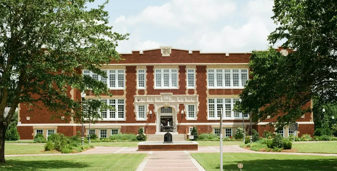 Stock photo of a red-brick school building seen across a green lawn, with large trees on both sides.